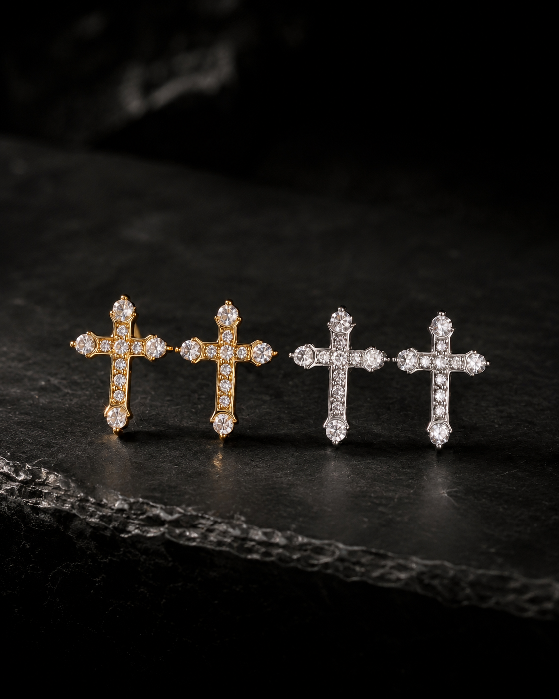 Four small cross-shaped jewelry pieces arranged in a row on a dark textured surface. Two are gold-toned with clear stones, and two are silver-toned with clear stones, set against a very dark background.